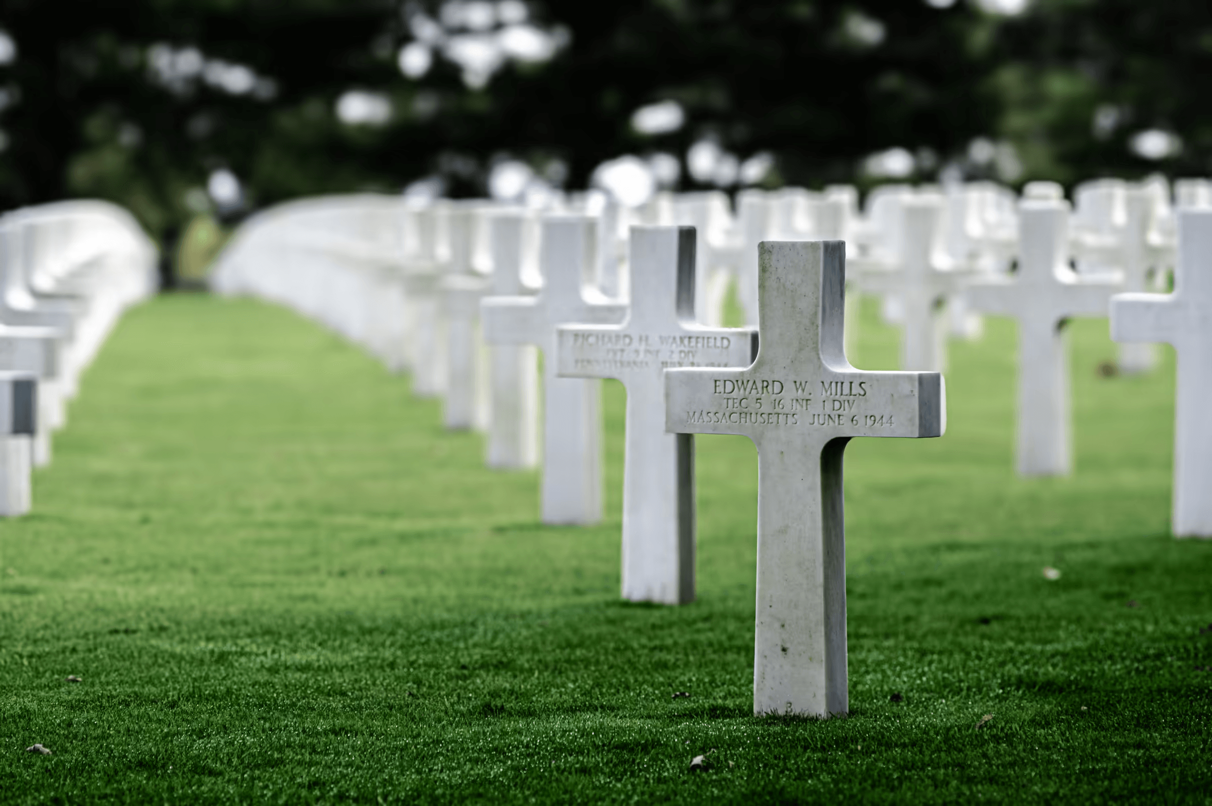 Landscape photograph of the Normandy American Cemetery and Memorial in Colleville-sur-Mer, France. Rows of white crosses stretch across a green lawn, with a close-up of one cross bearing the inscription 'EDWARD W. MILLS MASSACHUSETTS JUNE 6 1944'.  Trees line the background. The image conveys a sense of solemnity, remembrance, and respect.