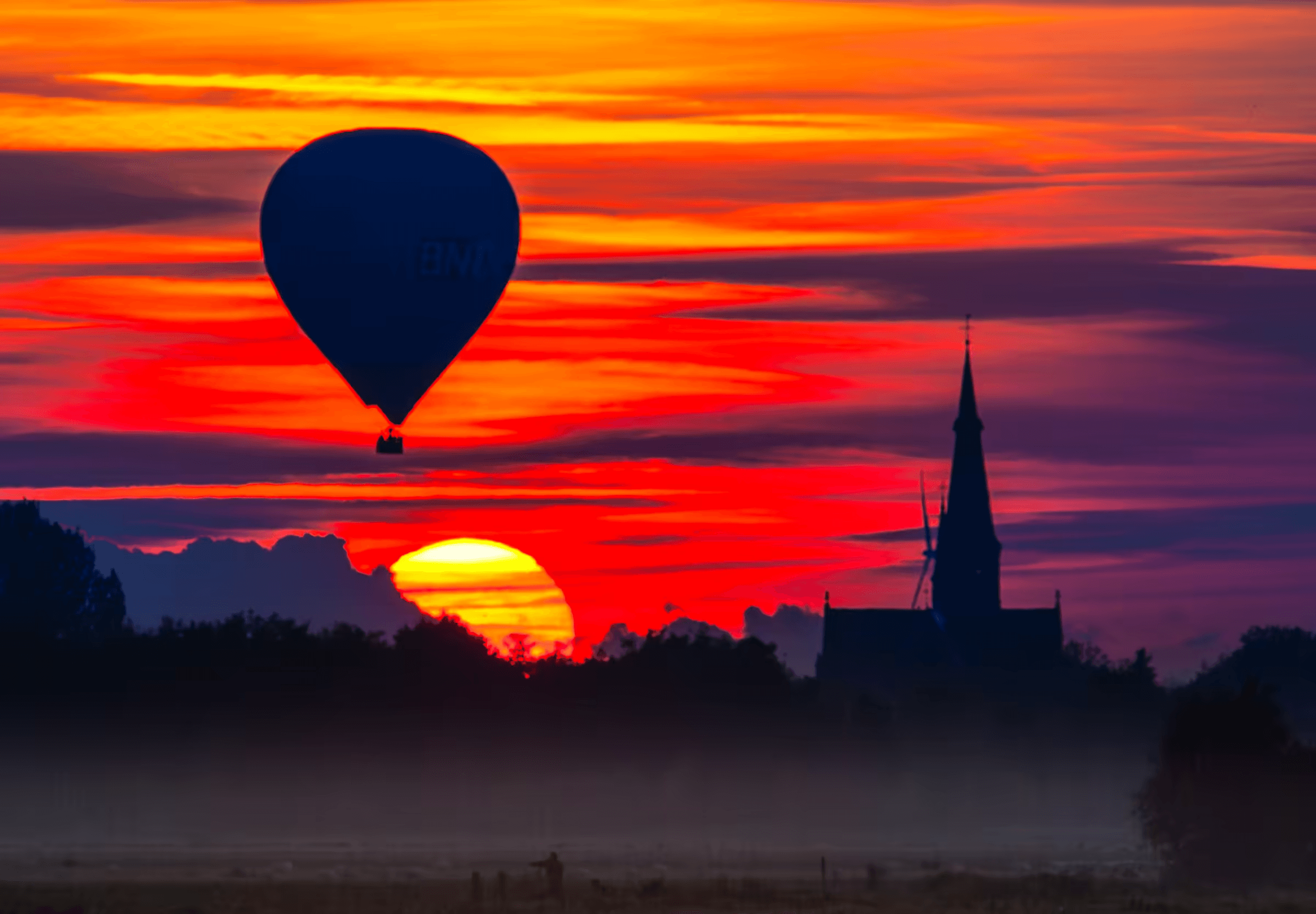 Landscape photograph of a hot air balloon silhouetted against a vibrant sunset in North Brabant, Netherlands. The dark silhouette of a hot air balloon drifts across a sky ablaze with orange, red, and purple sunset colors. A church spire and treeline are also silhouetted on the horizon.  Dramatic sunset landscape with hot air balloon, North Brabant, Dutch countryside.