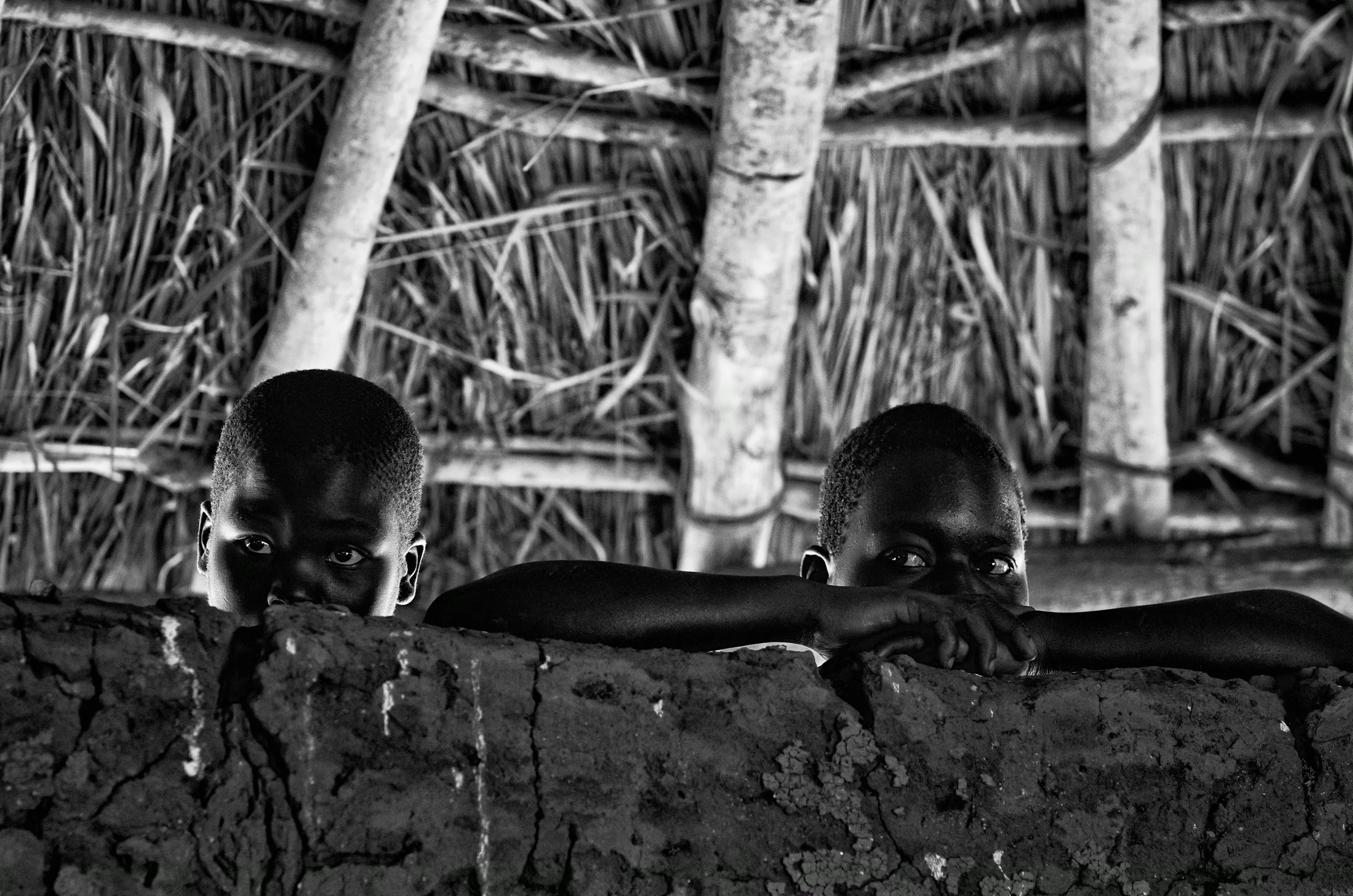 Black and white photograph of two young children in North Uganda peeking over a rough mud wall, looking directly at the camera, inside a traditional structure..