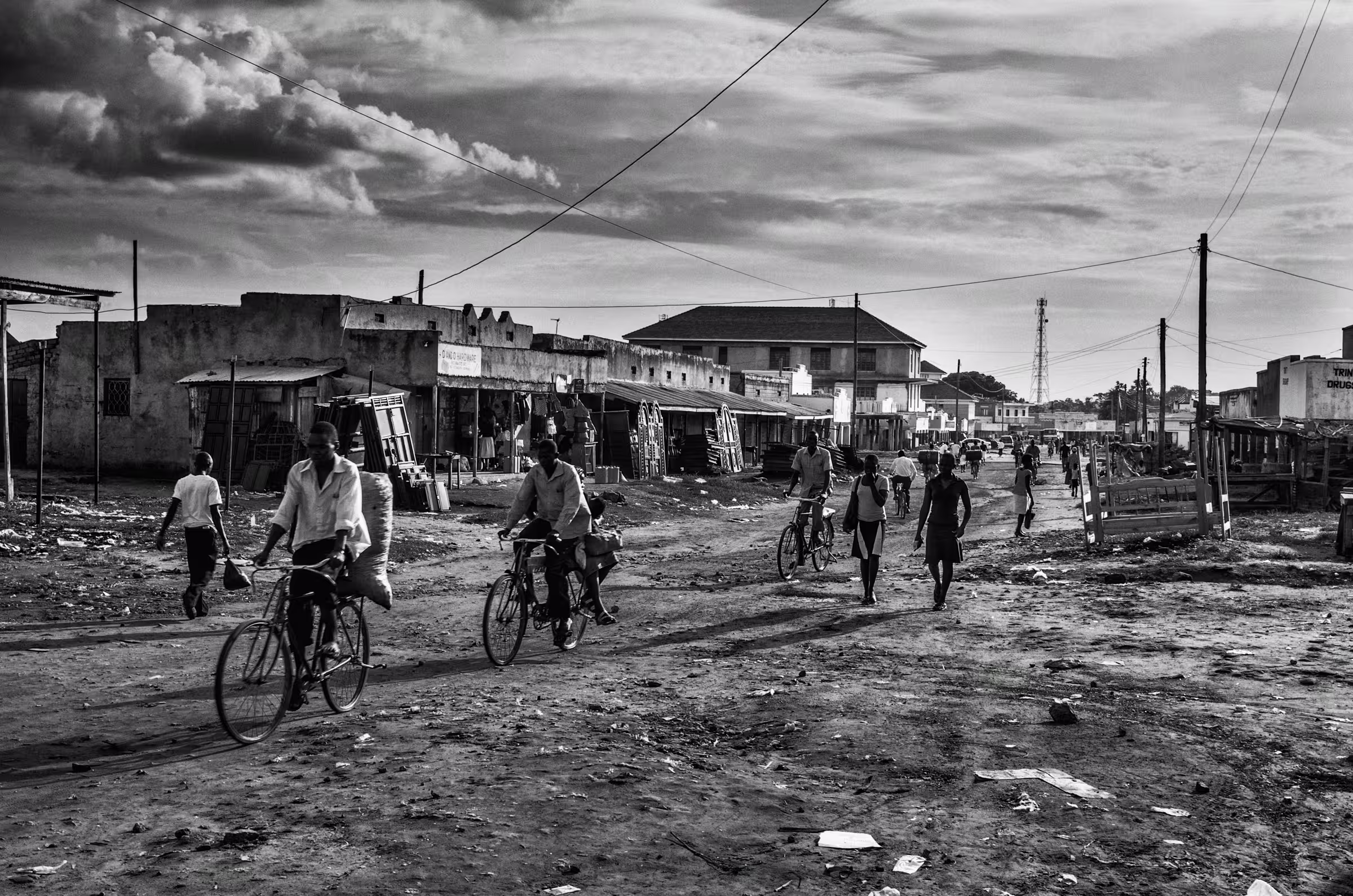Black and white street scene photograph in a refugee settlement in Northern Uganda. People are walking and cycling on a dirt road lined with simple buildings and shops.