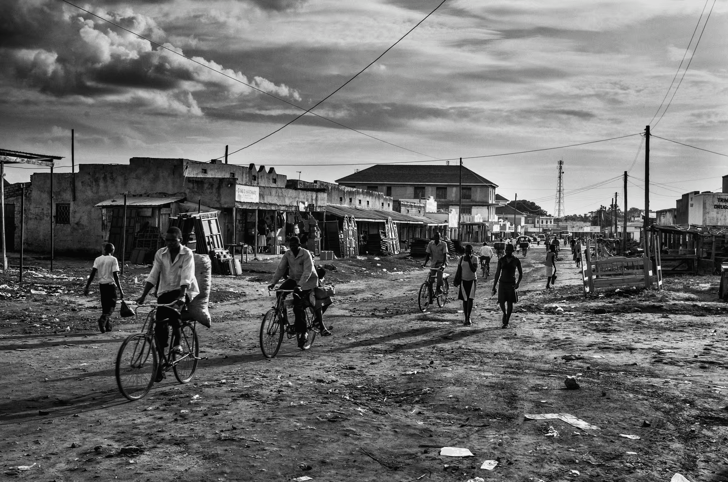 Black and white street scene photograph in a refugee settlement in Northern Uganda. People are walking and cycling on a dirt road lined with simple buildings and shops.