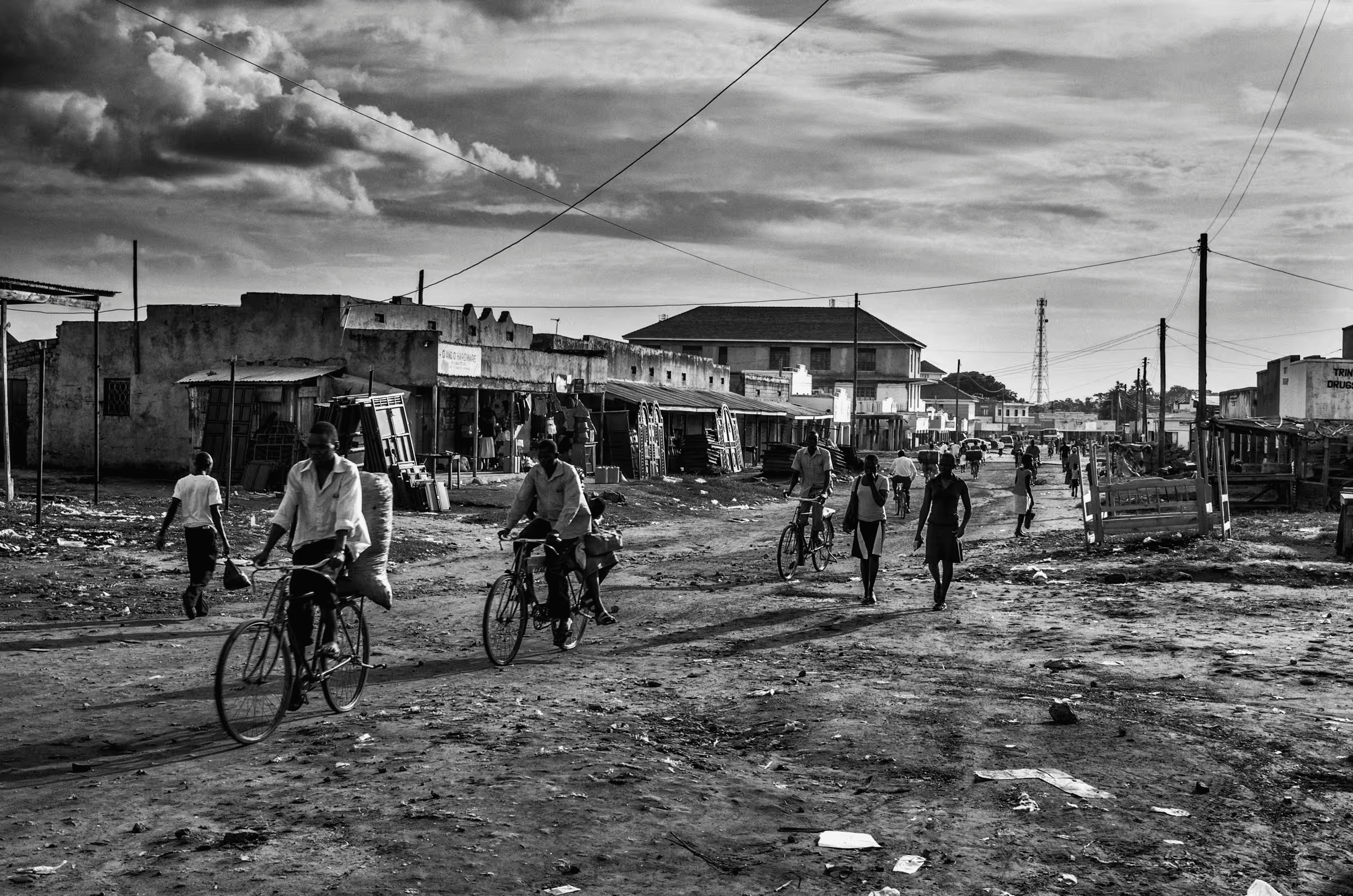 Black and white street scene photograph in a refugee settlement in Northern Uganda. People are walking and cycling on a dirt road lined with simple buildings and shops. The sky is cloudy and dramatic. The image captures a sense of everyday life and resilience in a challenging environment.