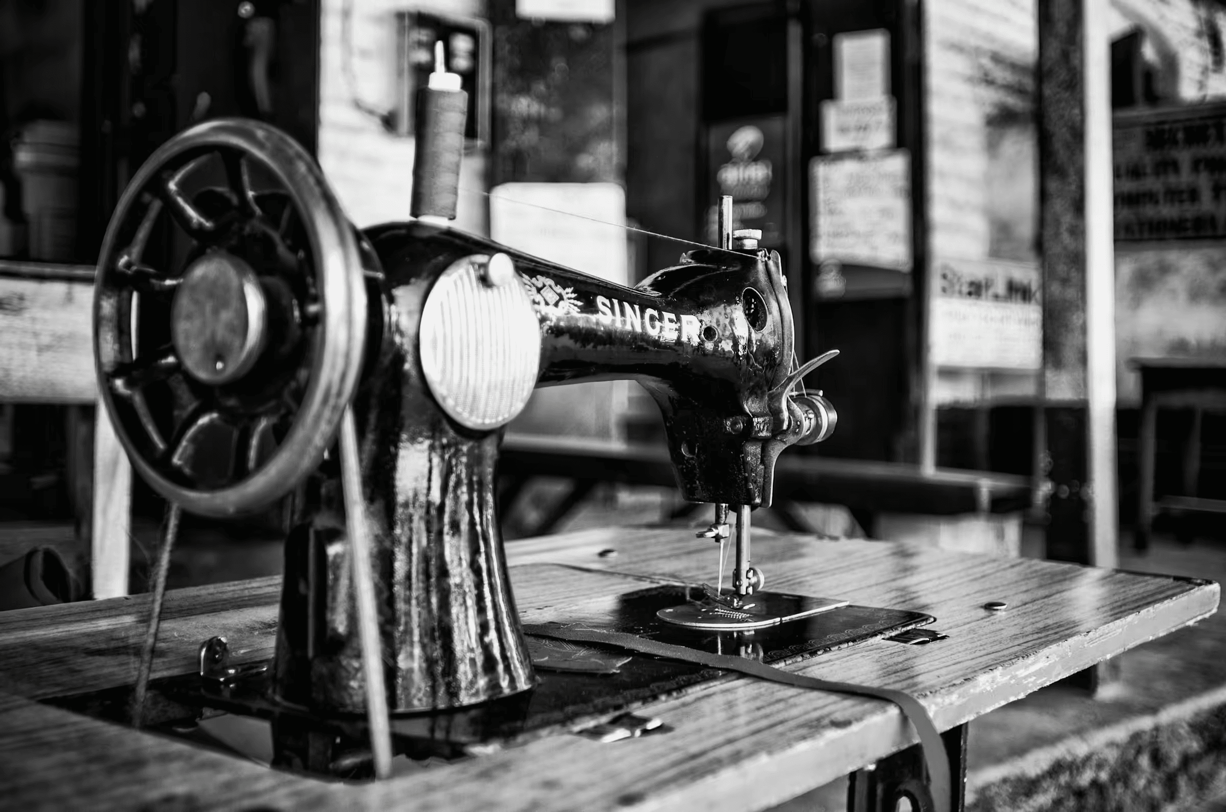 Black and white photograph of a vintage Singer sewing machine in Botswana. The machine, prominently displaying the 'SINGER' logo, sits on a wooden table. Details of the mechanical parts, wheel, and needle are visible. The background suggests a simple, possibly outdoor setting in Botswana. Evocative monochrome image of a classic sewing machine.