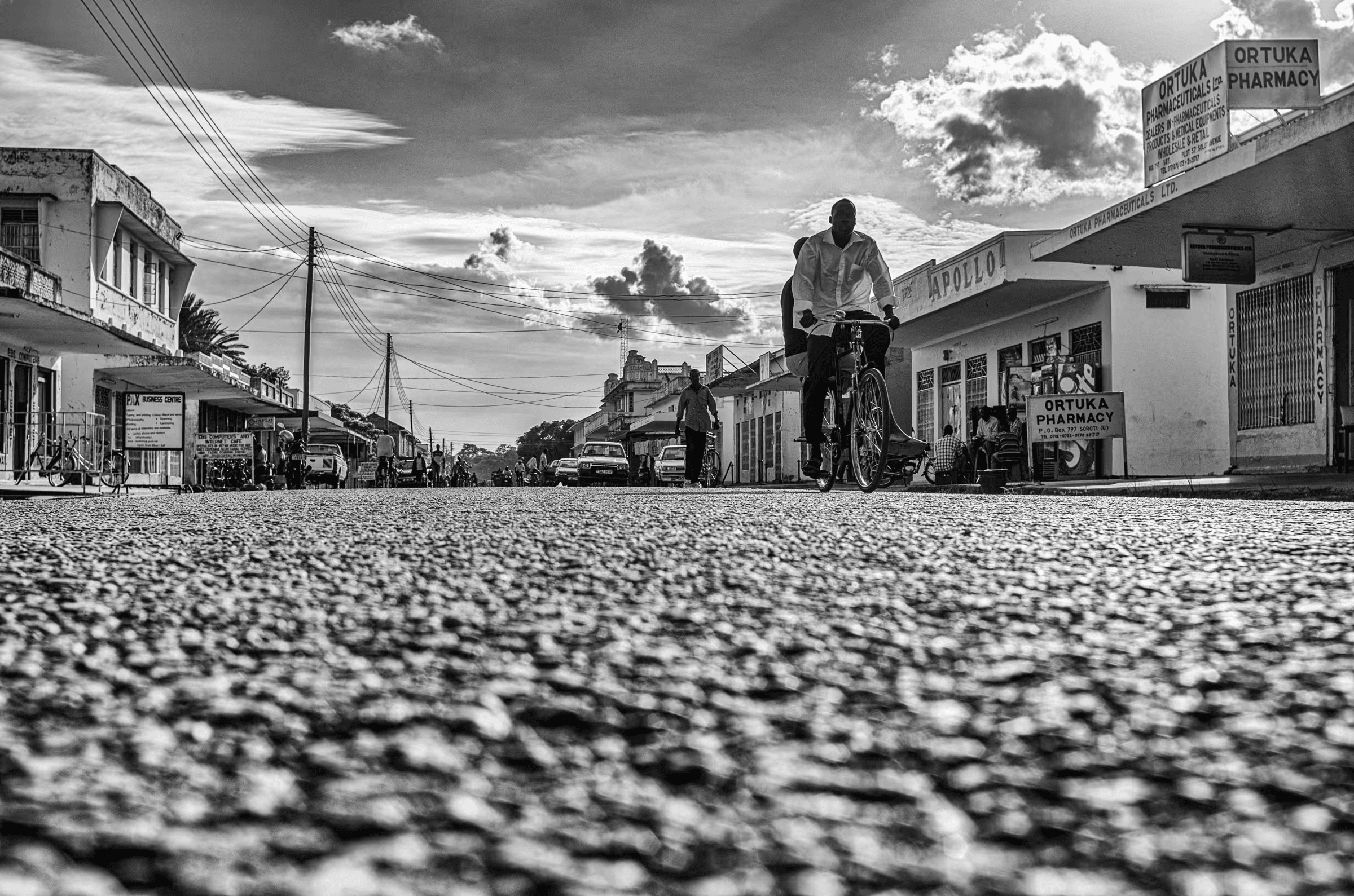 Black and white street scene photograph in Ortuka, Northern Uganda. People are walking and cycling on a dirt road. Buildings with signs for 'ORTUKA PHARMACY' and 'APOLLO' are visible. The sky is cloudy. The image captures a sense of daily life in a Ugandan town.