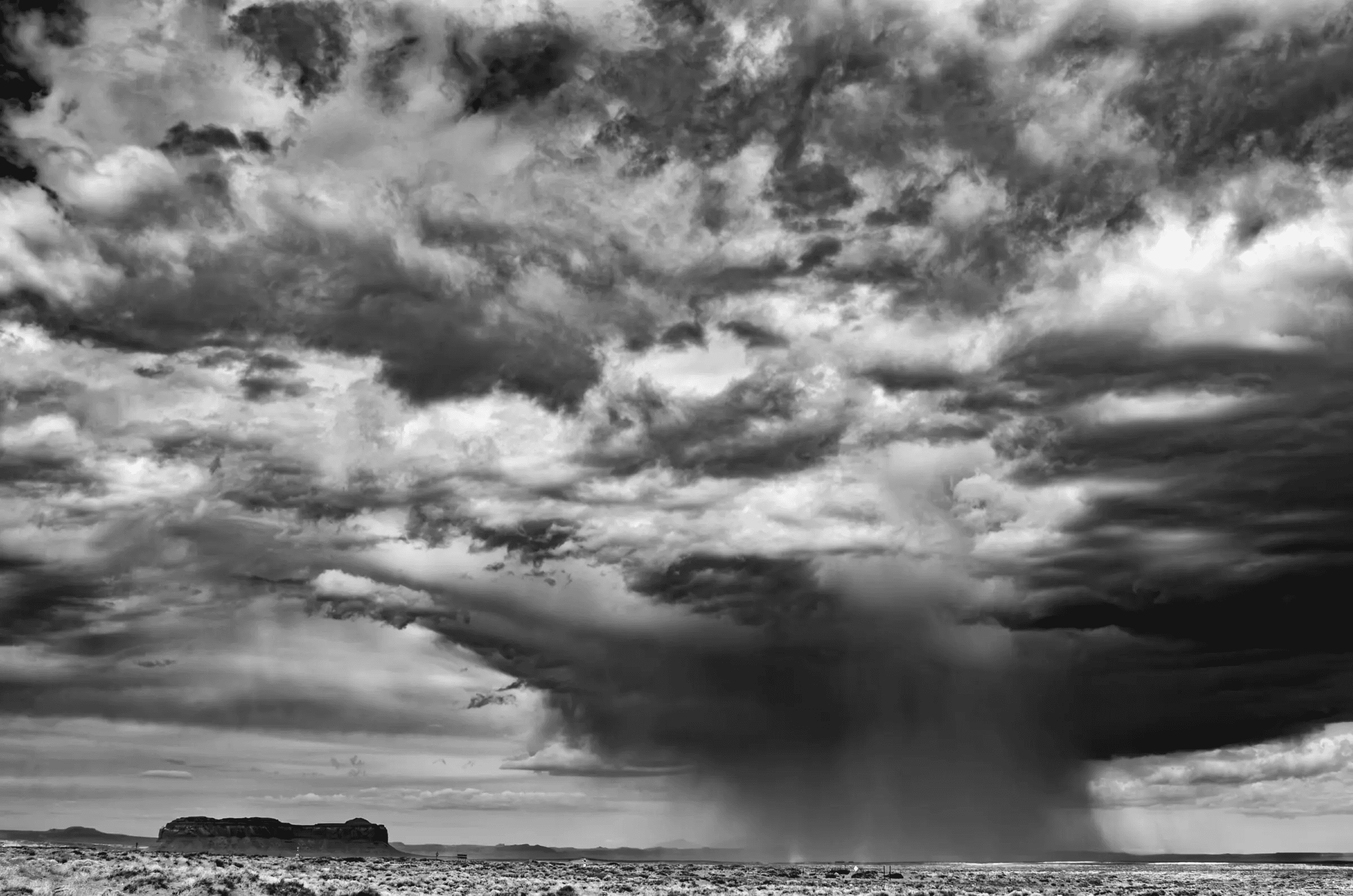 Rainfall, High Desert Black and white photograph of a dramatic sky over a flat desert landscape, with a heavy rain shaft falling from dark clouds near a distant mesa.