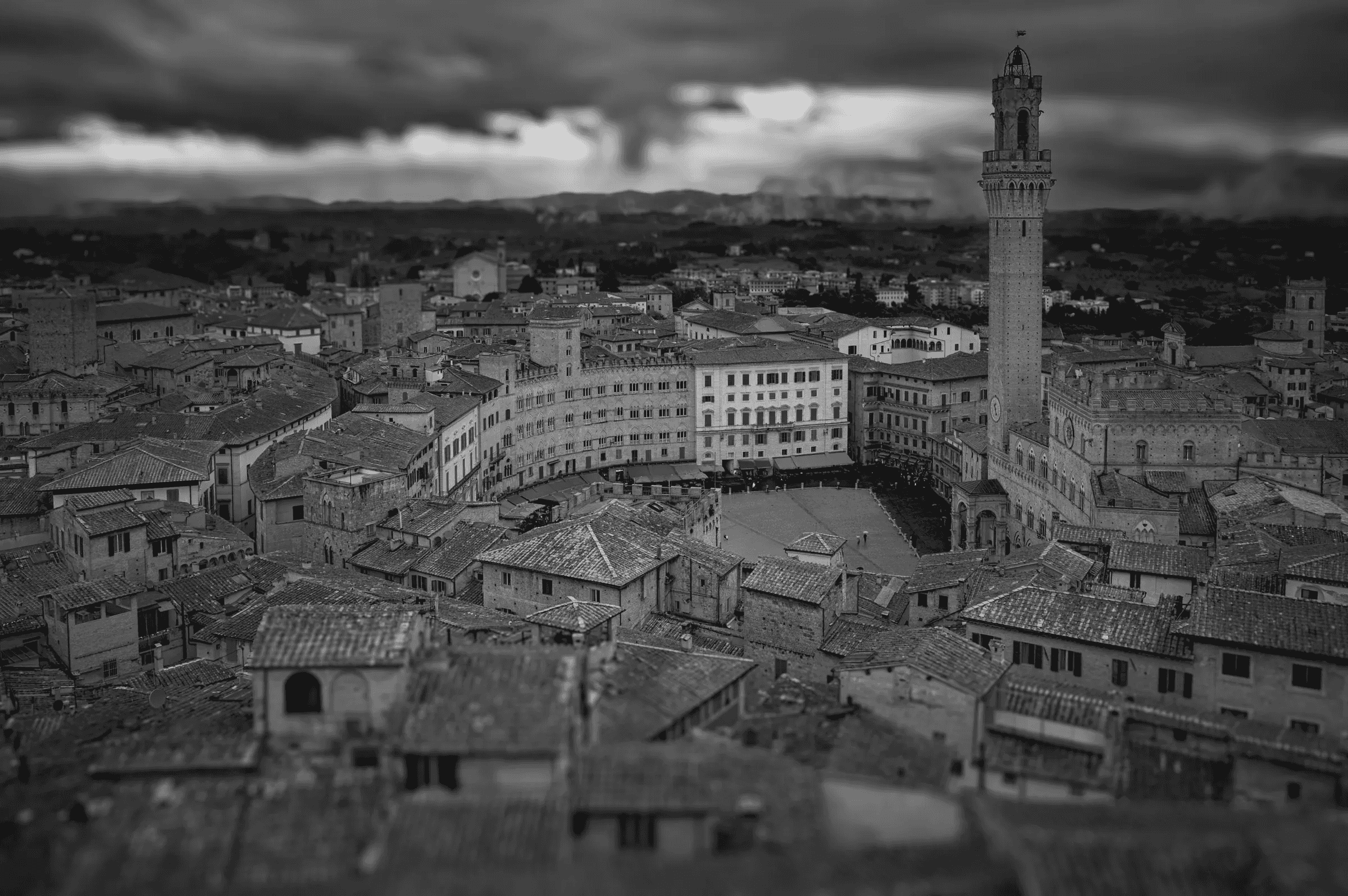 Black and white photograph taken from a high angle overlooking the Piazza del Campo in Siena, Italy, showing the distinctive curved piazza, surrounding buildings, and the Torre del Mangia under a dramatic sky.