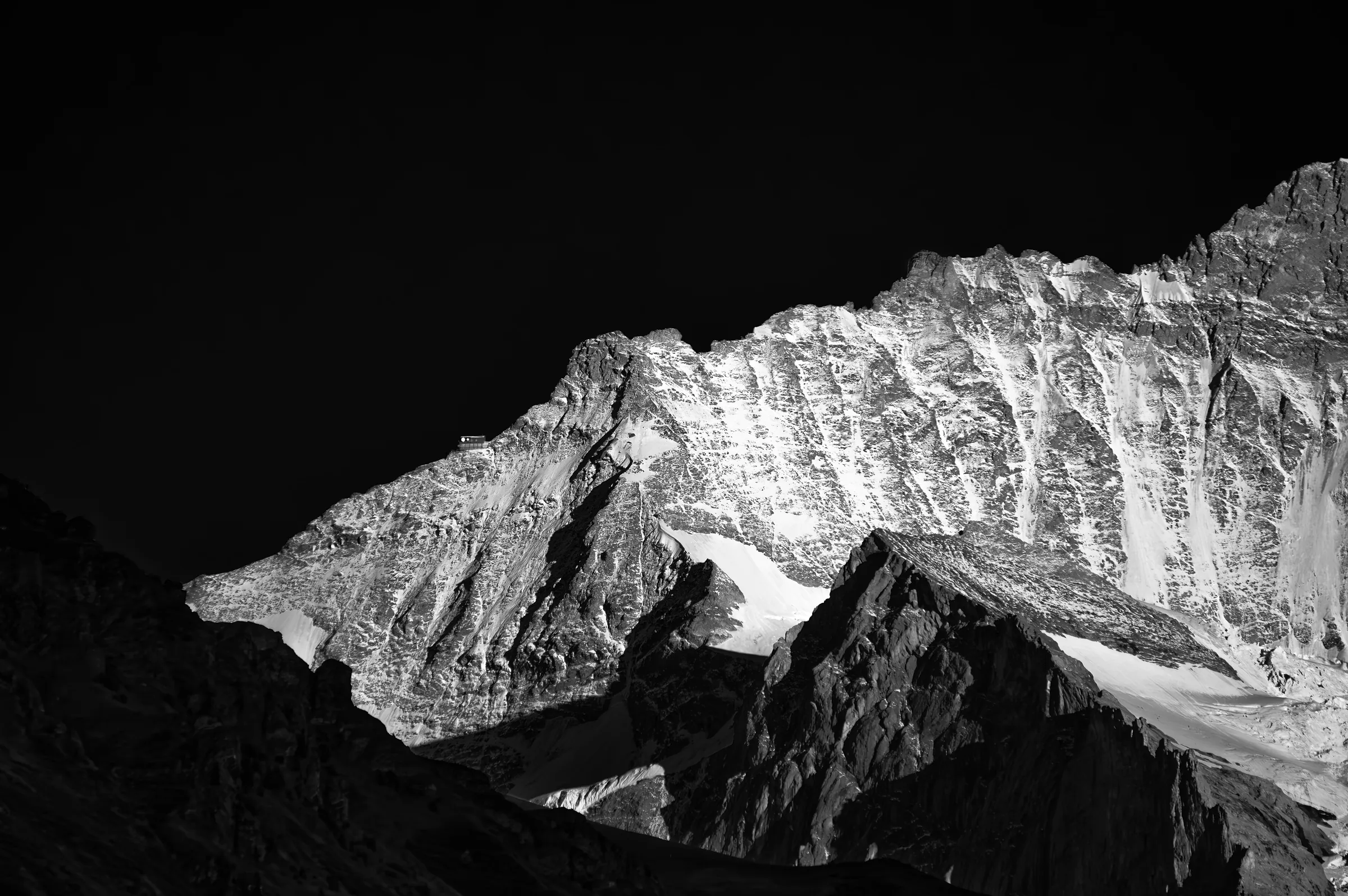 High-contrast black and white photograph showing sharp, snow-dusted rocky mountain peaks against a black sky in the Swiss Alps.