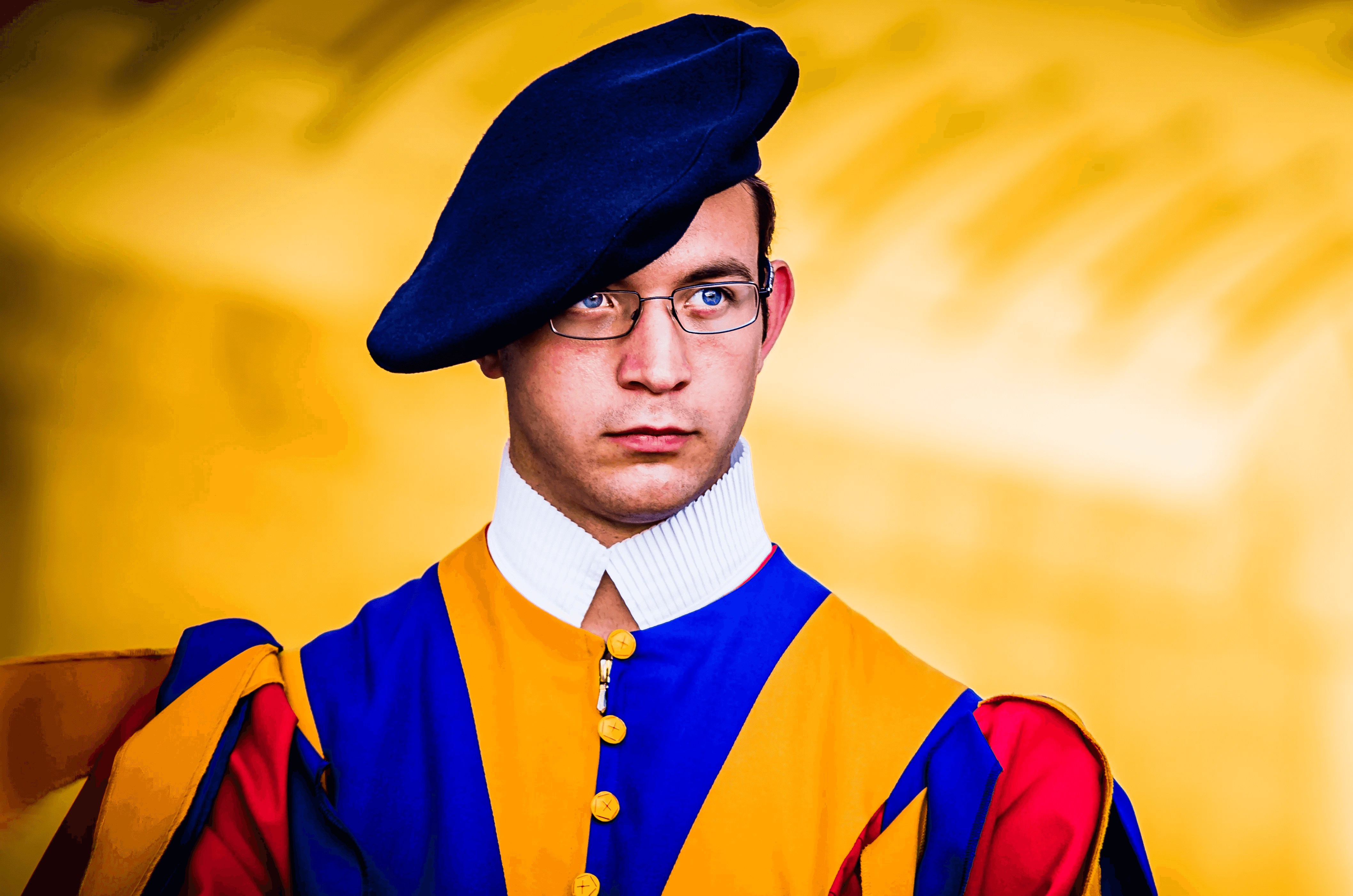 Close-up color portrait of a Swiss Guard at the Vatican. He wears the traditional colorful blue, yellow, and red Renaissance-style uniform, a dark blue beret, and glasses.