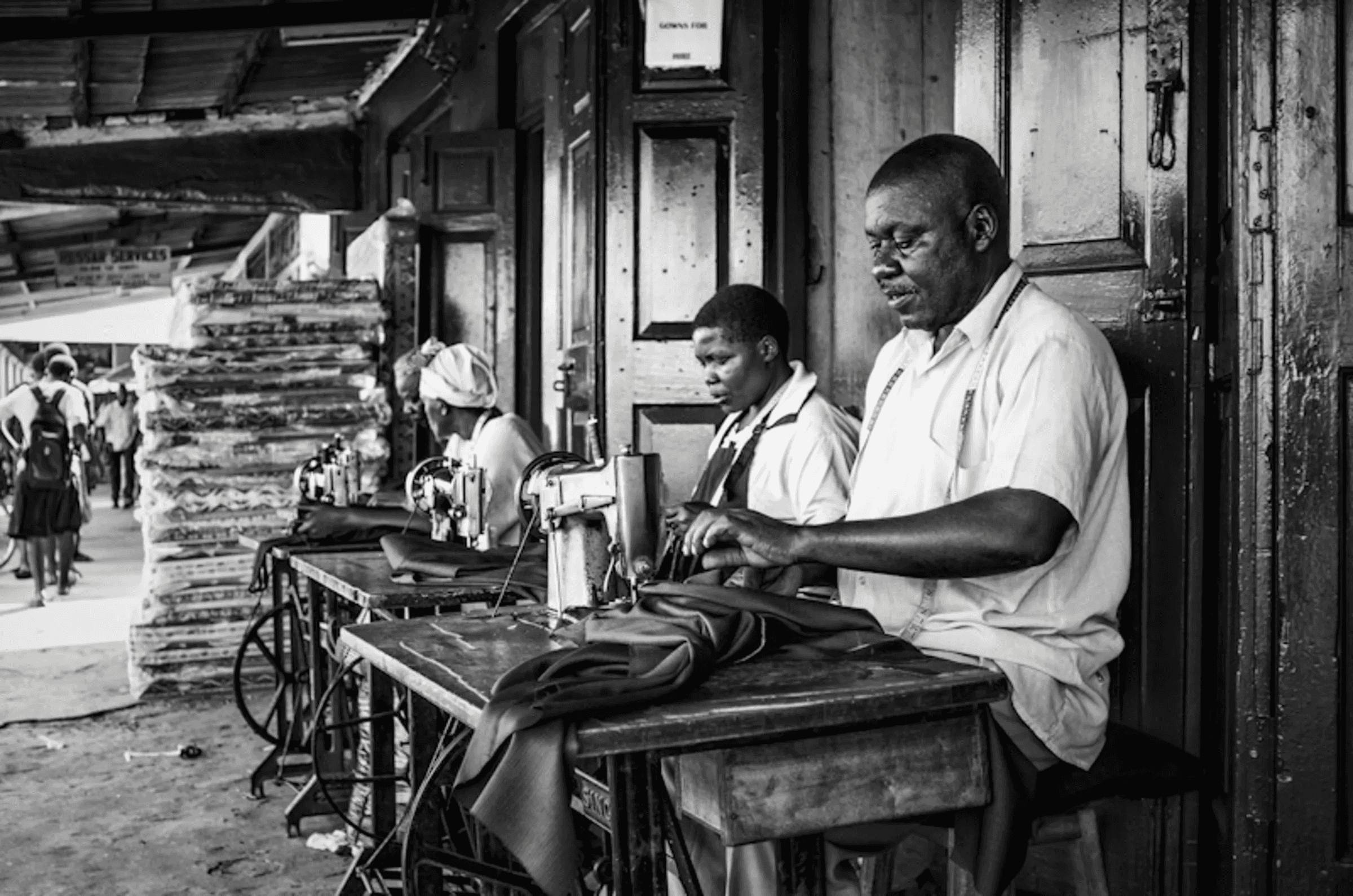 Black and white photograph of three people, two women and a man, working intently at sewing machines set up outdoors, possibly in a market or workshop.