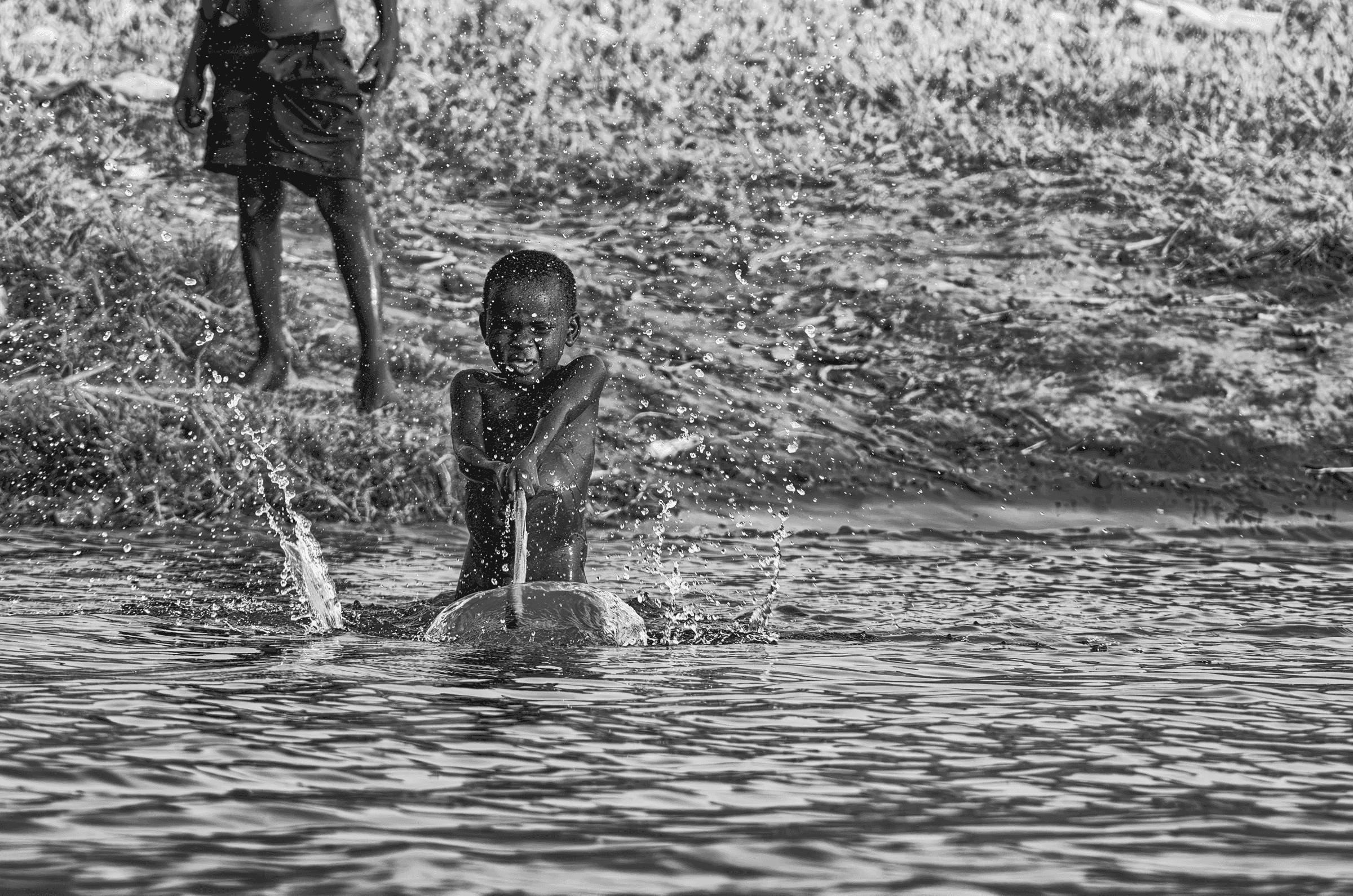 Black and white photograph of a young Ugandan child sitting in water near the bank and joyfully splashing water towards the camera.