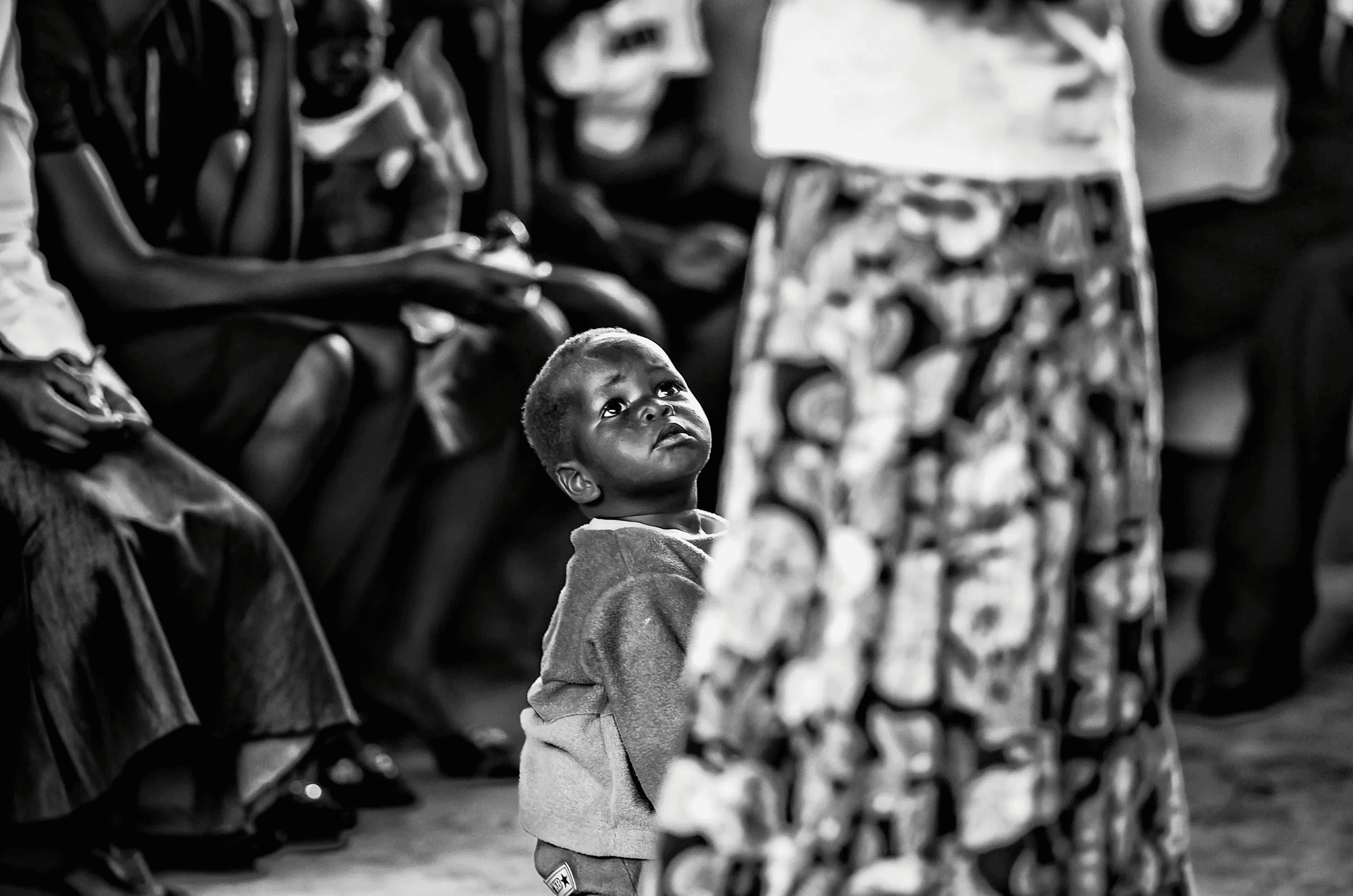 Black and white photograph focusing on a small Ugandan child looking upwards, partially obscured by an adult figure in the foreground.