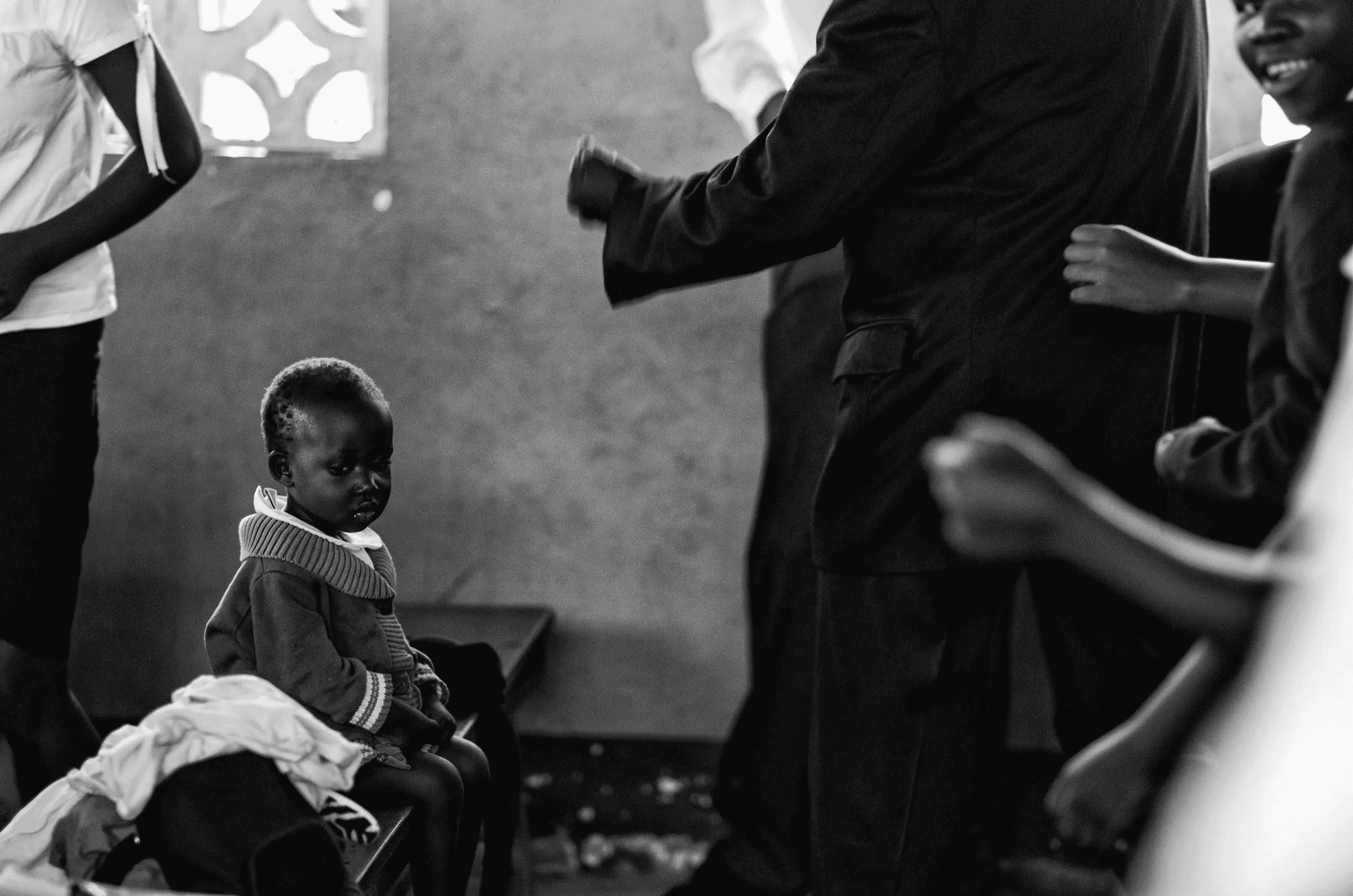 Black and white photograph of a small Ugandan child sitting quietly on a bench, observing activity in the background where other people are gathered indoors.