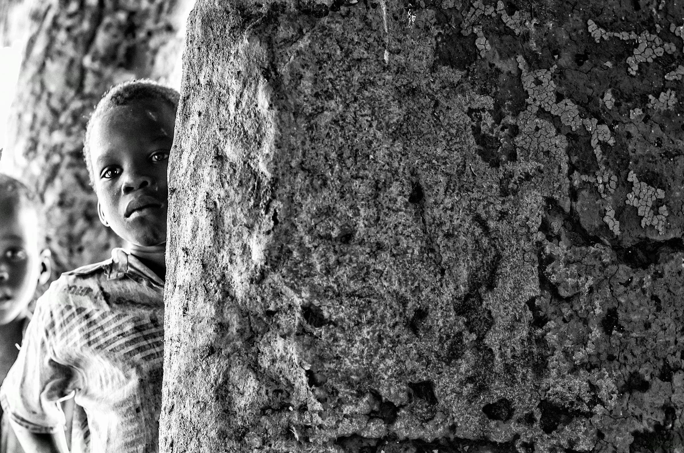 Black and white photograph of a young Ugandan child peeking around a rough, textured pillar or wall, looking towards the camera.