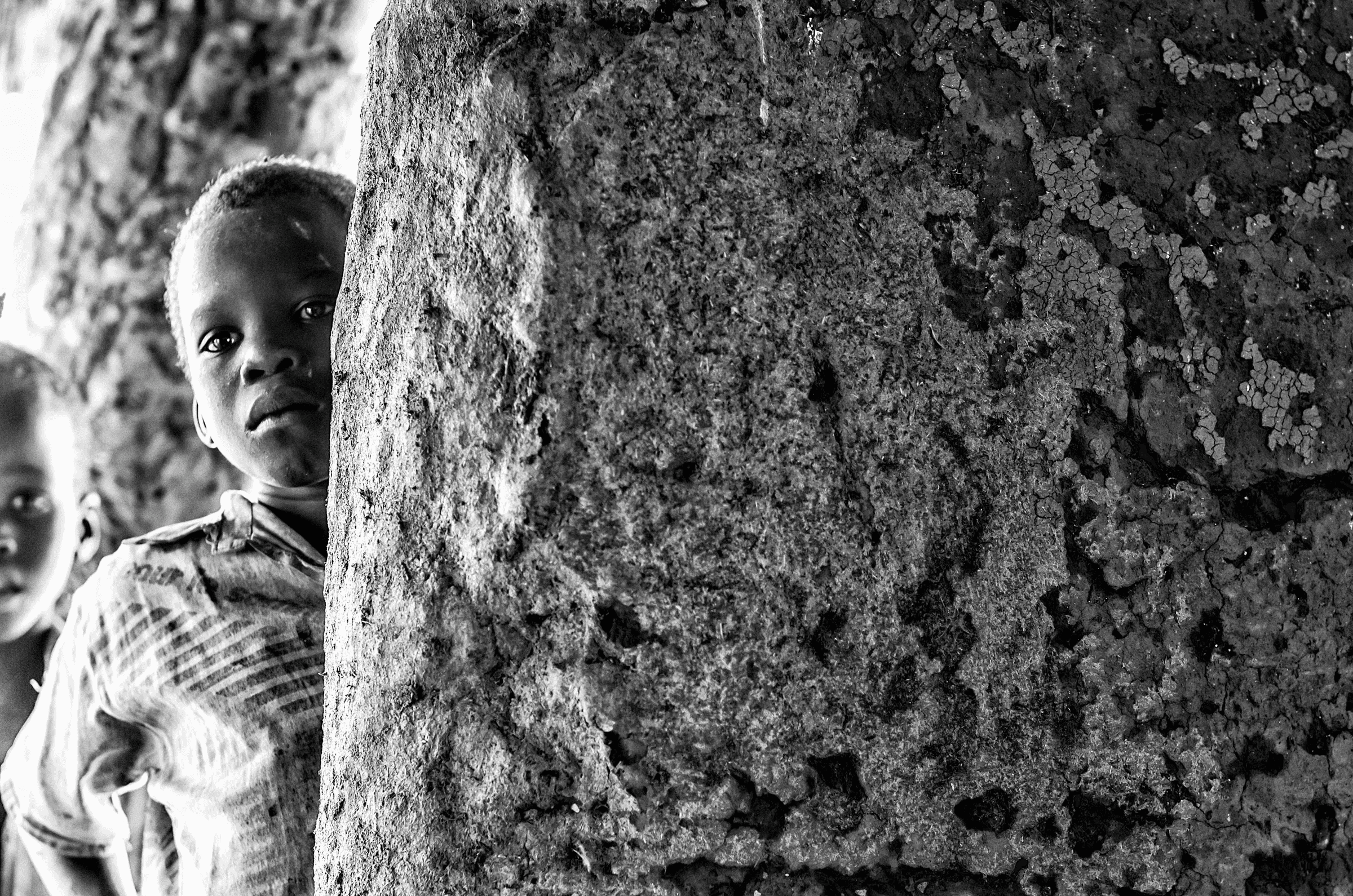 Black and white photograph of a young Ugandan child peeking around a rough, textured pillar or wall, looking towards the camera.