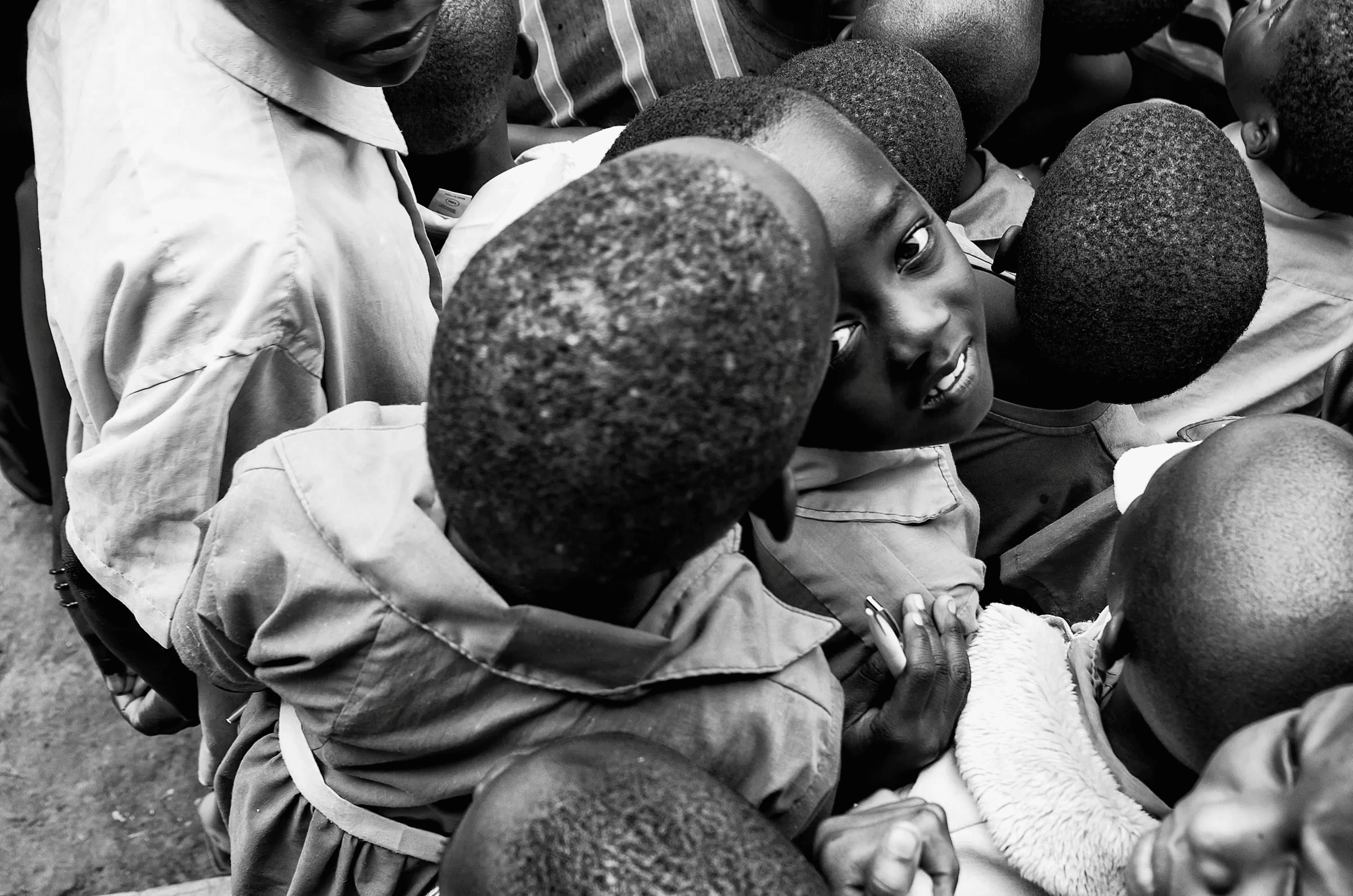 Black and white photograph taken from a high angle, looking down into a group of Ugandan children, one of whom looks directly up at the camera.