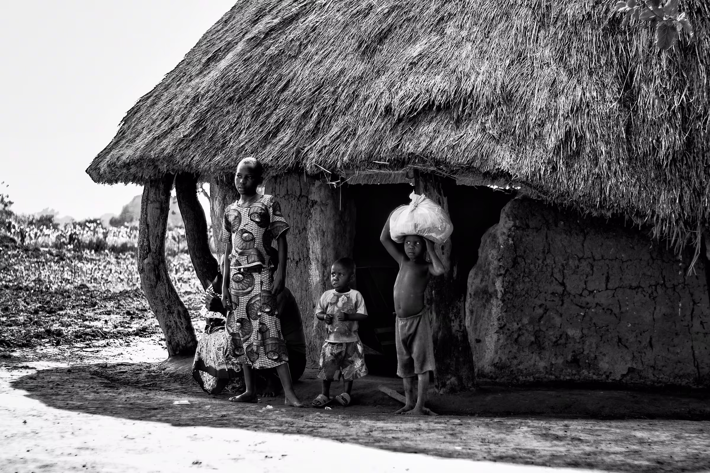 Black and white photograph of a Ugandan family standing outside their traditional mud hut with a thatched roof.