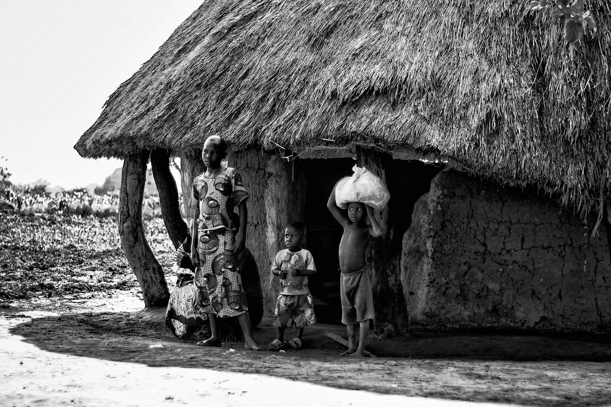 Black and white photograph of a Ugandan family standing outside their traditional mud hut with a thatched roof.  A woman stands beside two young children, one of whom carries a large bundle on their head.  The setting is a rural village, showcasing a glimpse into daily life in Uganda.