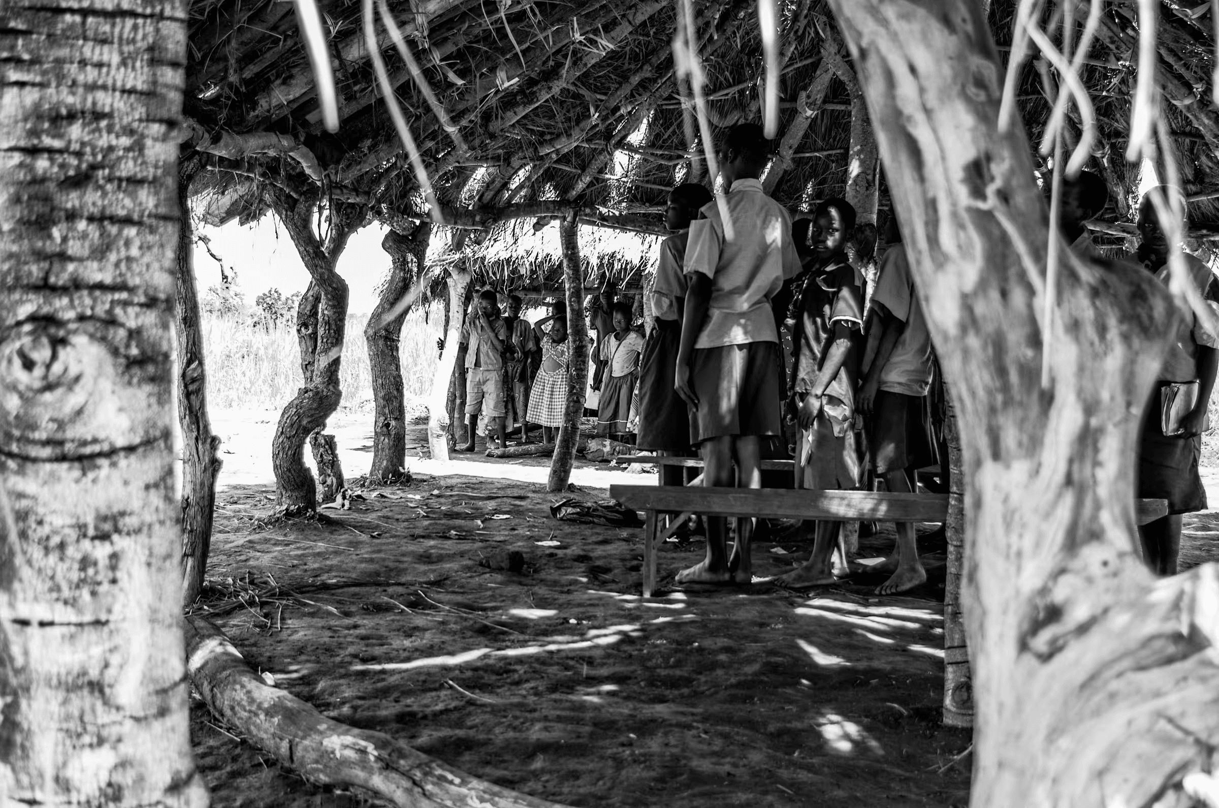 Black and white photograph showing a group of people, mostly children and some adults, gathered under a rustic thatched-roof structure in a rural Ugandan village. The scene is framed by tree trunks and branches. The image captures a sense of community and everyday life.