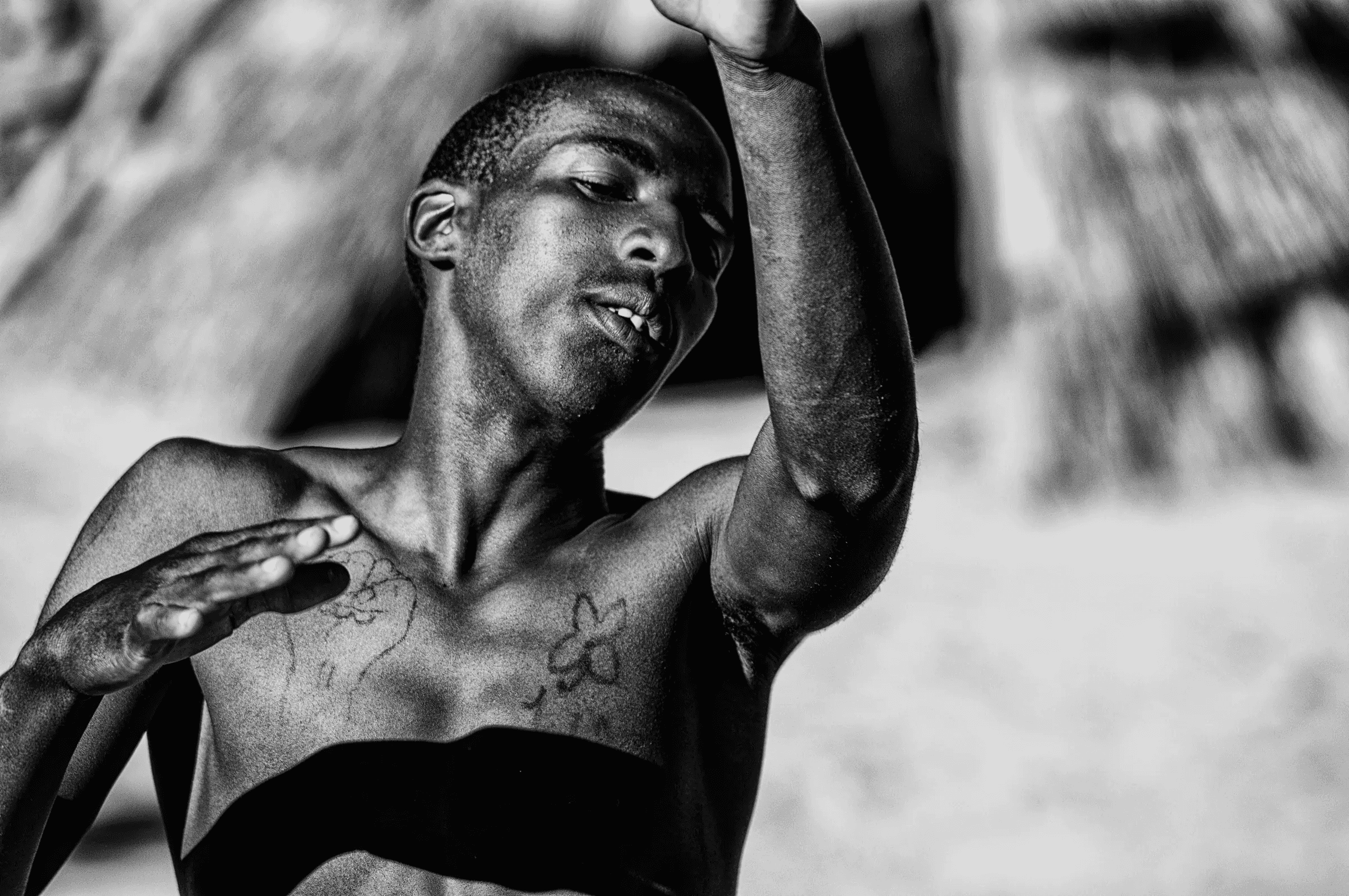 Black and white portrait of a shirtless young man from Vaalwater, South Africa, captured mid-movement with arms raised, showing chest tattoos in bright sunlight.