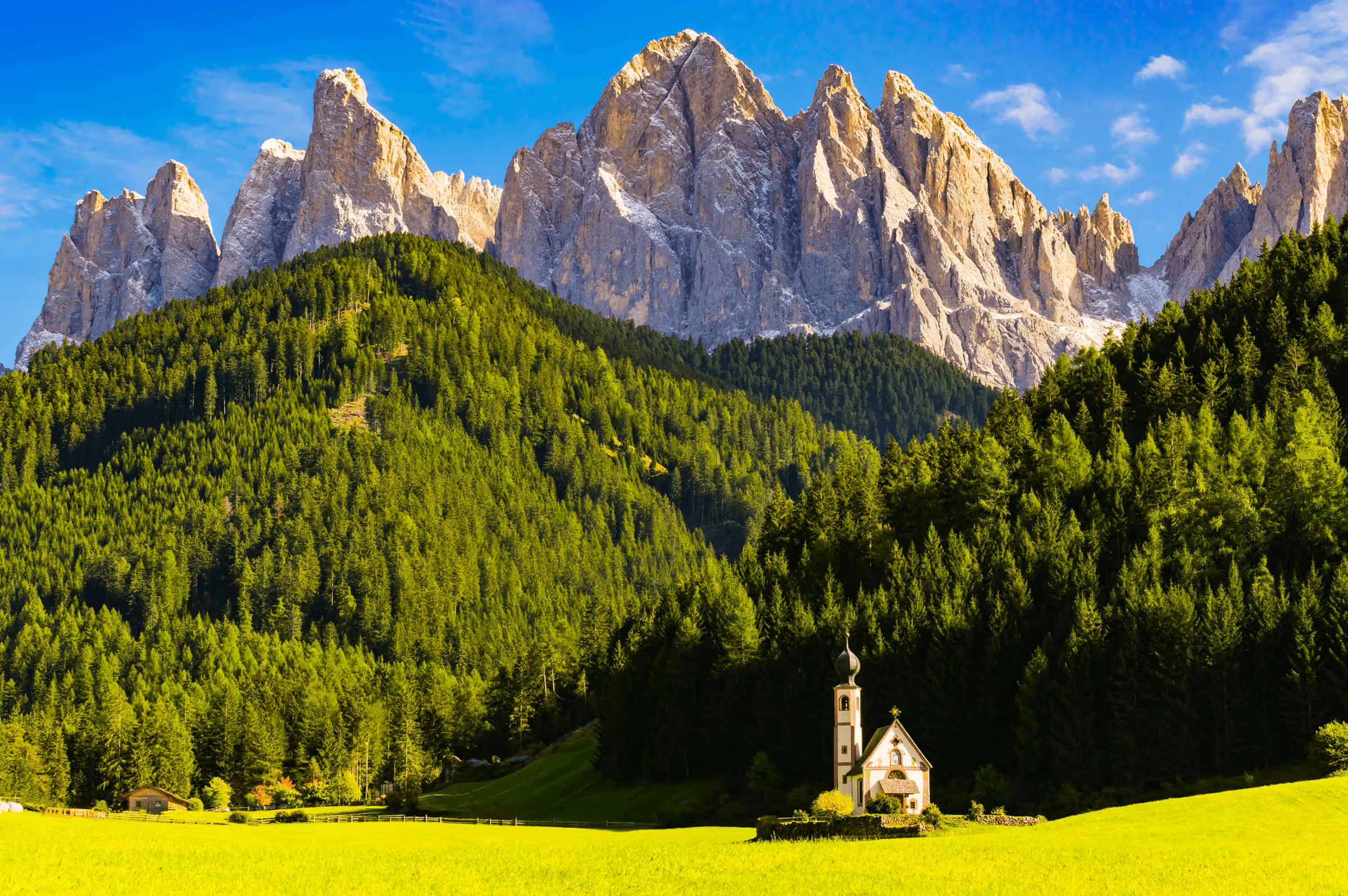 Landscape photograph of the Val di Funes in the Dolomites, Italy.  The iconic Church of St. John stands in a green meadow in the foreground.  Behind it, a dense forest rises up the slopes of a mountain.  The dramatic, jagged peaks of the Odle Group dominate the background under a clear blue sky.