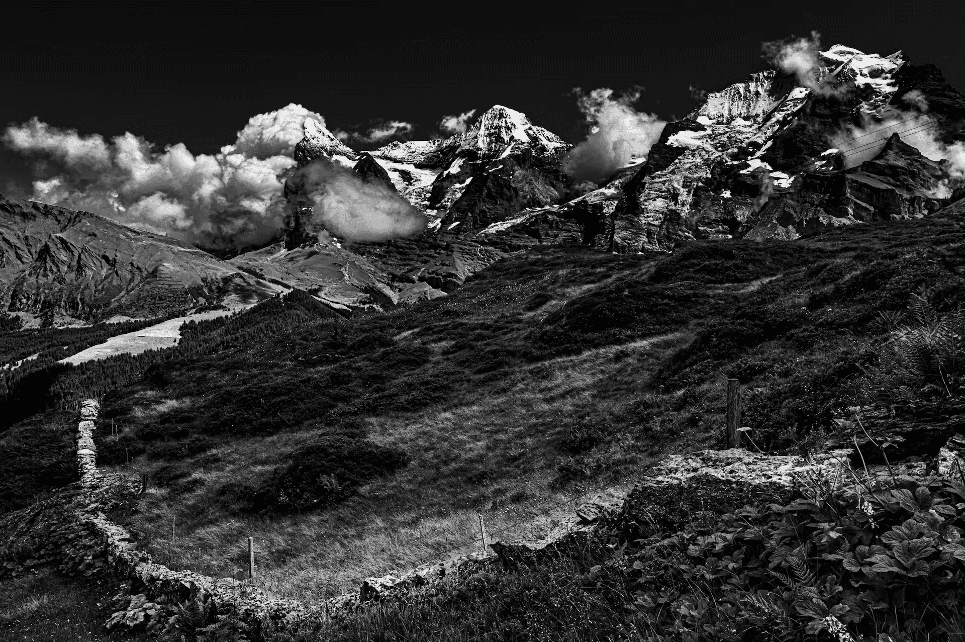 A black and white landscape photograph featuring a rustic stone wall in the foreground leading the eye towards the majestic Eiger, Mönch, and Jungfrau mountains in the Swiss Alps.