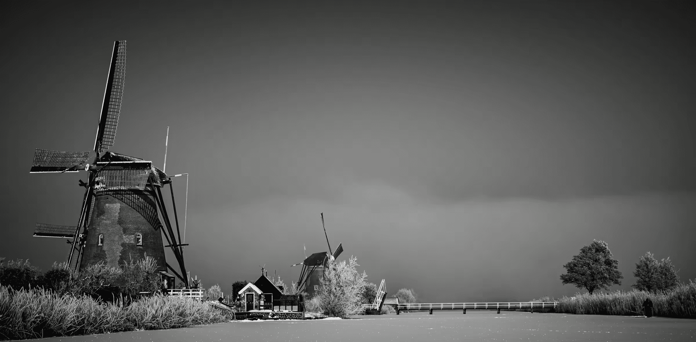 Black and white landscape photograph of windmills at Kinderdijk, Netherlands. Silhouetted windmills stand against a misty sky, with a small bridge and traditional Dutch architecture visible in the mid-ground.