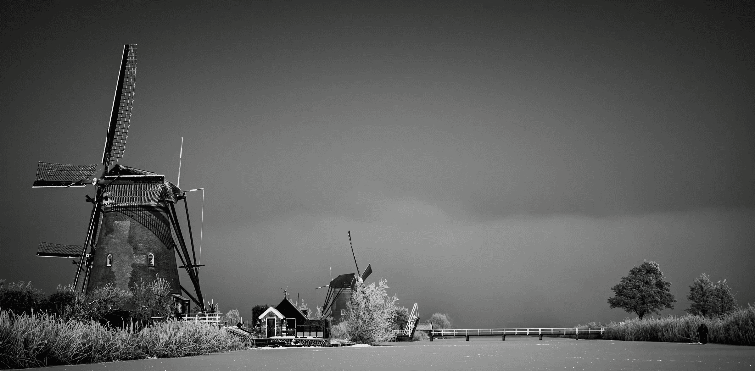 Black and white landscape photograph of windmills at Kinderdijk, Netherlands. Silhouetted windmills stand against a misty sky, with a small bridge and traditional Dutch architecture visible in the mid-ground.