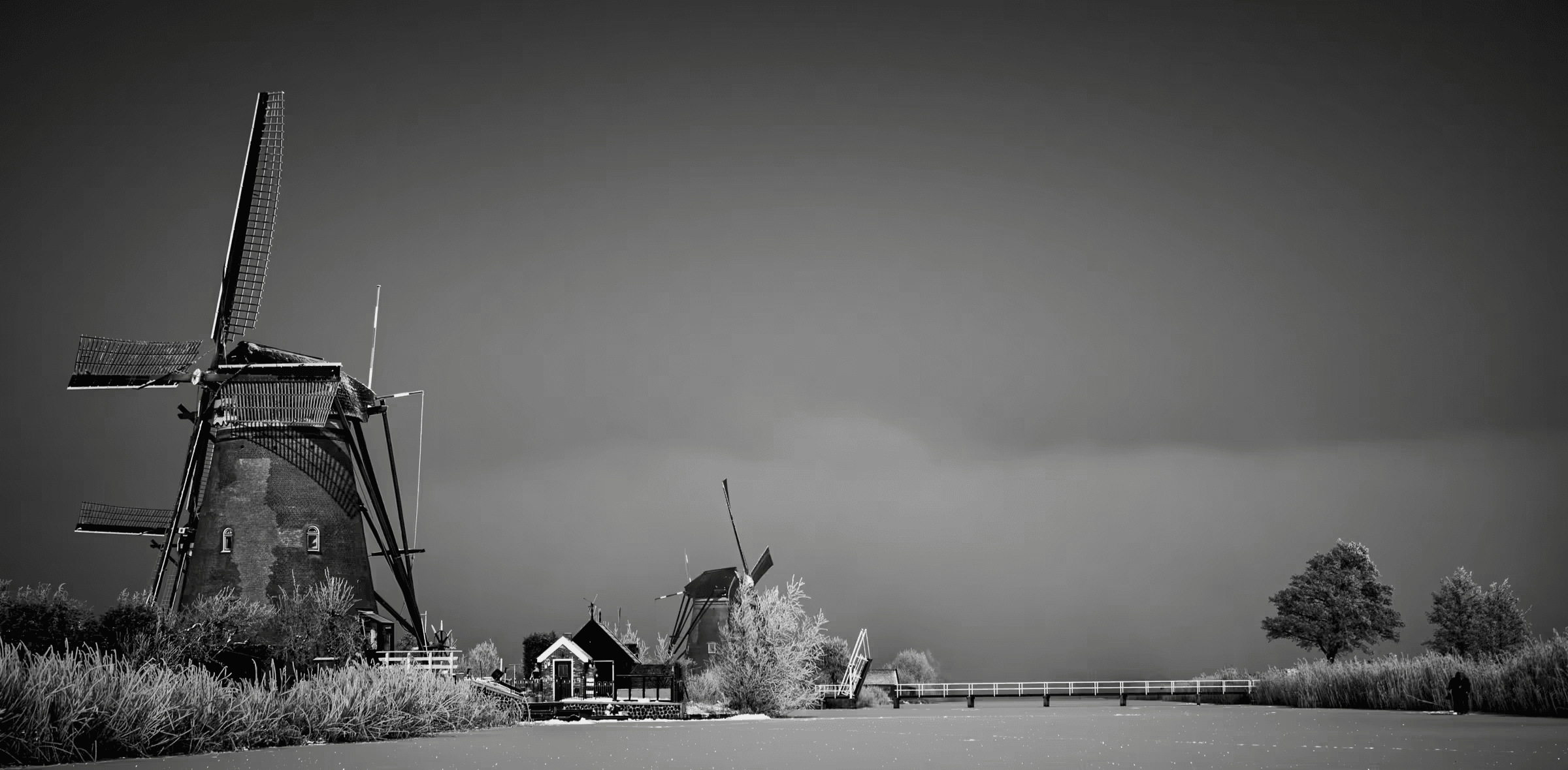 Black and white landscape photograph of windmills at Kinderdijk, Netherlands. Silhouetted windmills stand against a misty sky, with a small bridge and traditional Dutch architecture visible in the mid-ground. The image is rendered in monochrome tones, emphasizing the shapes and lines of the windmills and landscape. Evocative monochrome Dutch landscape photography.