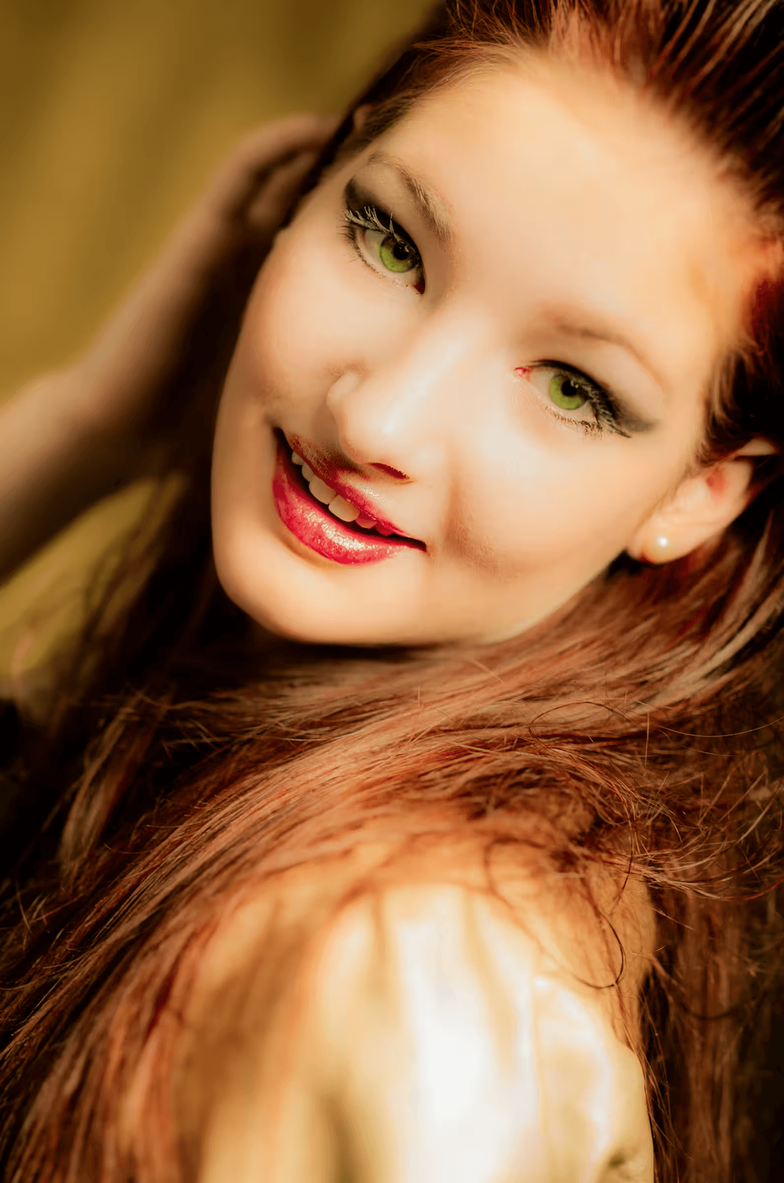 Portrait of a young woman with long, flowing auburn hair and striking green eyes. She is smiling slightly and looking towards the camera. Her makeup enhances her eyes. The background is a soft, out-of-focus warm tone.