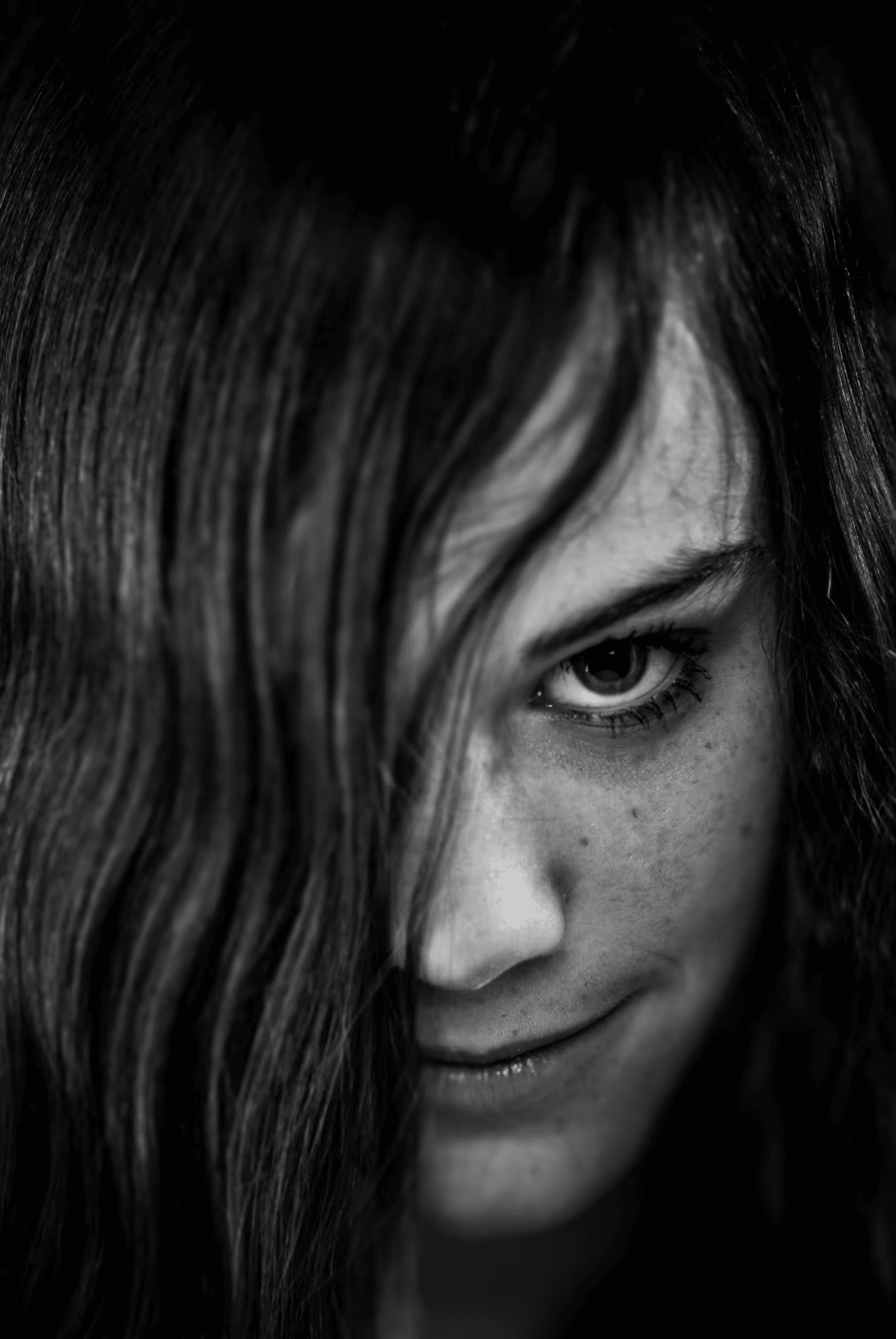 Black and white portrait of a woman with long hair partially covering her face. One eye is clearly visible and looking towards the camera, with a slight smile on her lips. The image emphasizes texture and contrast.