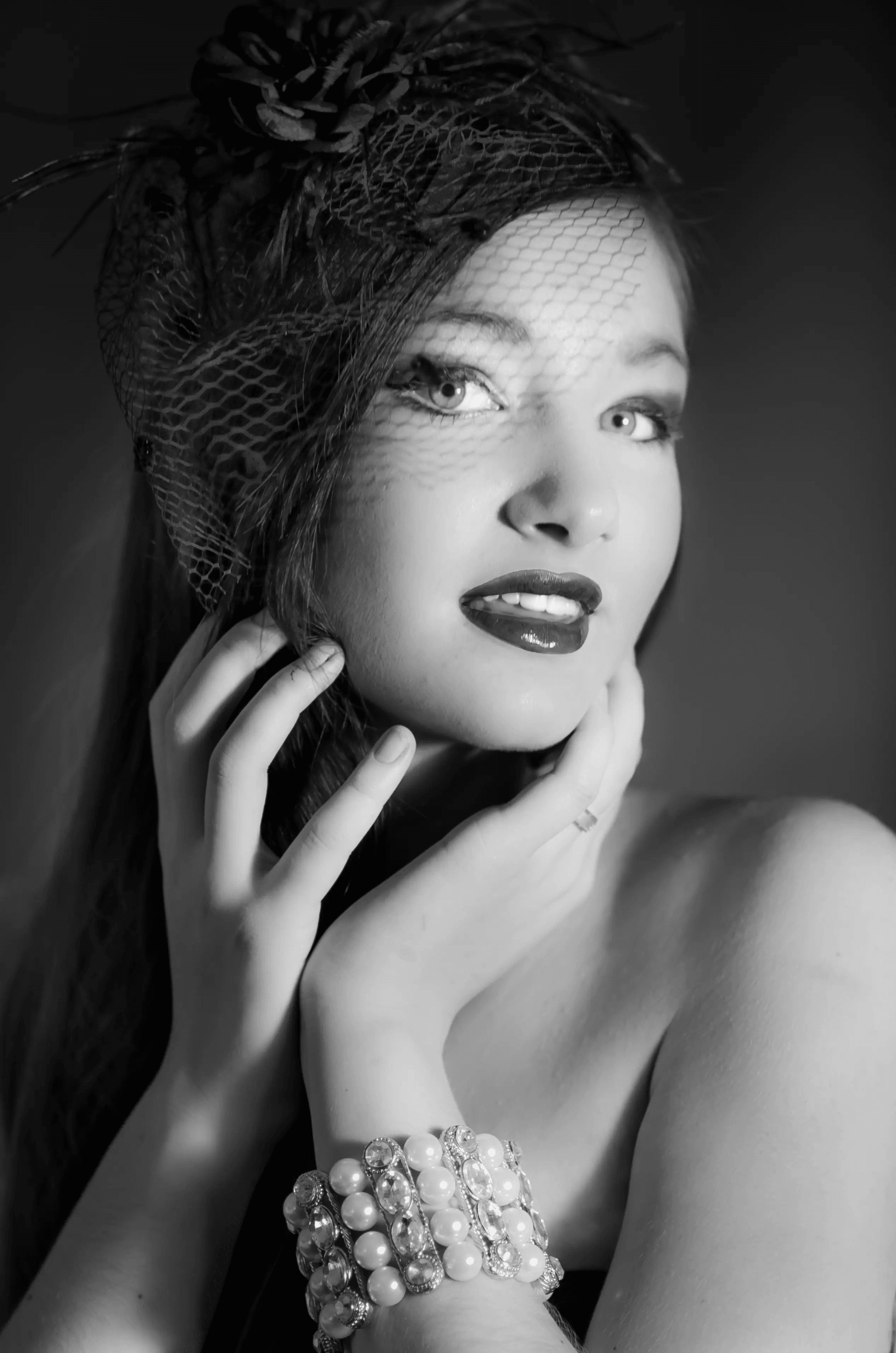 Black and white portrait of a woman wearing a vintage-style veiled headpiece and a pearl bracelet. She is looking slightly to the side with a soft smile. The image evokes a classic, glamorous Hollywood style.